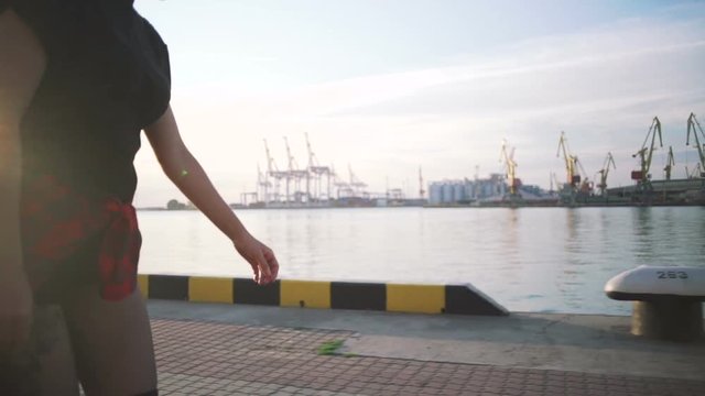 Young stylish funky girl with green hair riding roller skates and dancing near sea port during sunset