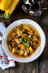 Soup of yellow zucchini with potatoes, carrots, onions, mushrooms, parsley and bacon on an old wooden background.