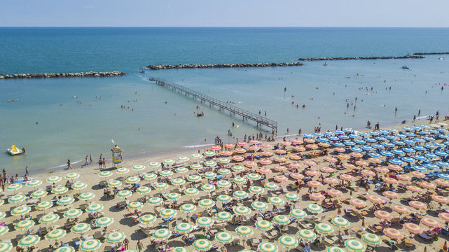 Dalla riviera romagnola vista aerea di ombrelloni colorati disposti in fila sulla spiaggia di Riccione. le persone in spiaggia prendono il sole o si riparano all' ombra prima di un bagno a mare.