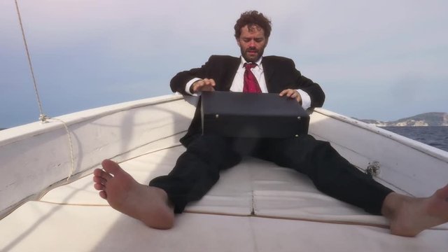 Man in suit sitting on a boat in the middle of the ocean opens his briefcase and works with his laptop
