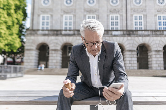 Senior businessman with ice cream cone sitting on bench looking at smartphone