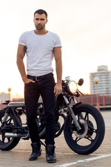 Naklejka premium Close up of a handsome rider guy in white blank t-shirt look to camera near classic style cafe racer motorcycle at sunset. Bike custom made in vintage garage. Brutal urban lifestyle. Outdoor portrait.