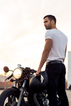 Sporty biker handsome rider guy in white blank t-shirt walk to classic style cafe racer motorbike on rooftop at sunset. Vintage bike custom made in garage. Brutal urban lifestyle. Outdoor portrait.