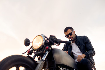 Handsome rider male with beard and mustache in black biker jacket, white shirt and fashion sunglasses smoke cigaret and sit on classic style cafe racer motorcycle at sunset. Brutal urban lifestyle.