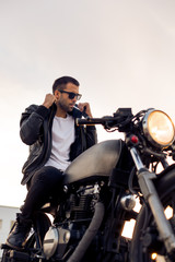 Fototapeta na wymiar Handsome young rider man with beard and mustache in black fashion sunglasses smoking cigaret and correct biker jacket sit on classic style cafe racer motorbike at sunset. Brutal fun urban lifestyle.