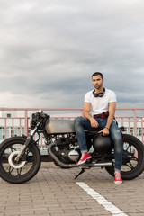 Fototapeta na wymiar Handsome rider guy with a beard and mustache sit on classic style biker cafe racer motorcycle and ready for long ride. Bike custom made in vintage garage. Brutal fun urban lifestyle. Outdoor portrait.
