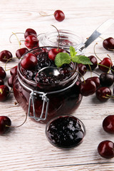 Jar of cherry jam, sour cherries and spoon on white wooden backg