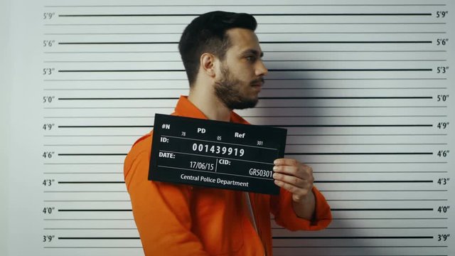 In a Police Station Arrested Man Getting Side, Front-View Mug Shot. He's in a Prisoner Orange Jumpsuit and Holds Placard. Height Chart in the Background. Shot on RED EPIC-W 8K Helium Cinema Camera.