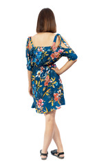 back view of standing young beautiful  woman.  girl  watching. Rear view people collection.  backside view of person. A girl in a flowered dress is standing with her hands on her hips.