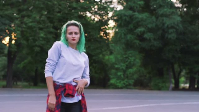 Young stylish funky girl with green hair riding roller skates and dancing in park, slow motion