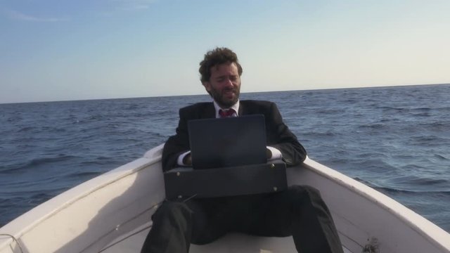 Man in suit sitting on a boat works with a laptop, gets hit by a wave and tries to save his computer