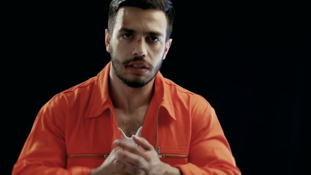 Aggressive Man with a Heavily Bruised Face Makes Threatening Gestures. He Wears Orange Prisoner Jumpsuit. Background is Isolated Black. Shot on RED EPIC-W 8K Helium Cinema Camera.