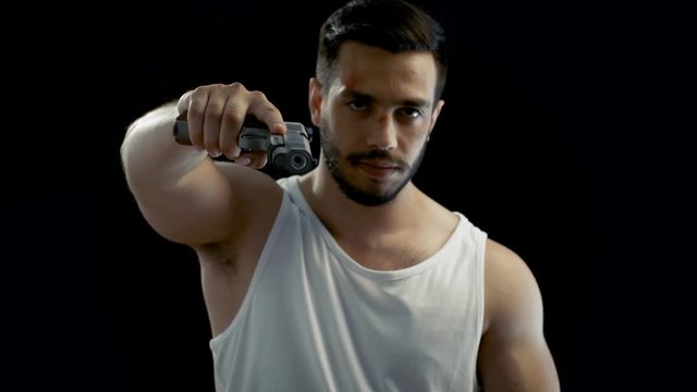 Aggressive Man with a Bruised Face Wearing Singlet, Aims His Gun into Camera and Threatens with it. Background is Isolated Black, Shot on RED EPIC-W 8K Helium Cinema Camera.