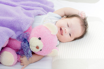 Portrait of a little adorable infant baby girl lying on back on the bed with bear doll and looking in camera indoors