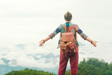 Hipster traveller with backpack and raised hands enjoying view at mountains in fog.