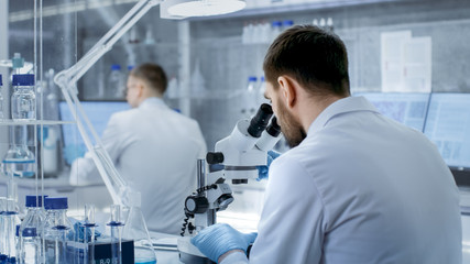 Shot of Research Scientist Adjusts His Microscope. He's Working in a High-End Modern Laboratory with Beakers, Glassware, Microscope and Working Monitors Surround Him.