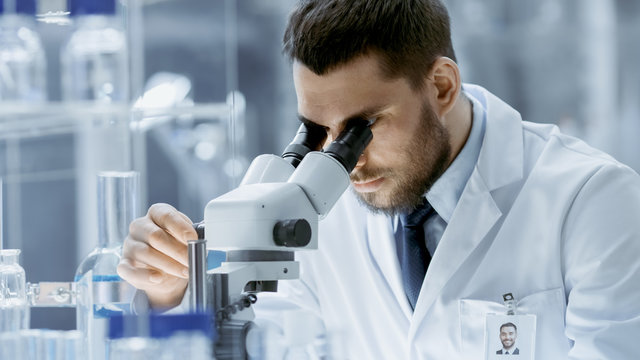 Close-up Shot of Research Scientist Adjusts His Microscope. He's Working in a High-End Modern Laboratory with Beakers, Glassware, Microscope and Working Monitors Surround Him.