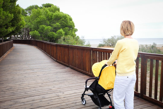 Young mother strolling a baby in carriage outdoors. Back view. Seashore with green trees on background