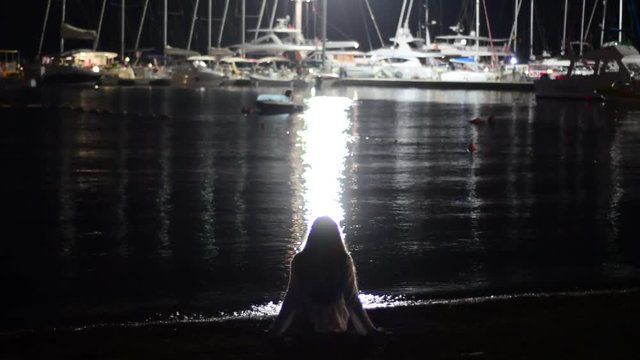 The girl sits in the evening on the seashore in the light of the lanterns. There are yachts in the distance.
