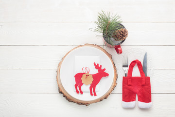 Christmas table setting. White plate with white card, deer figure decor, fork, knife, red mug with green twig decor on white wooden table with copy space. Christmas dinner concept. Flat lay