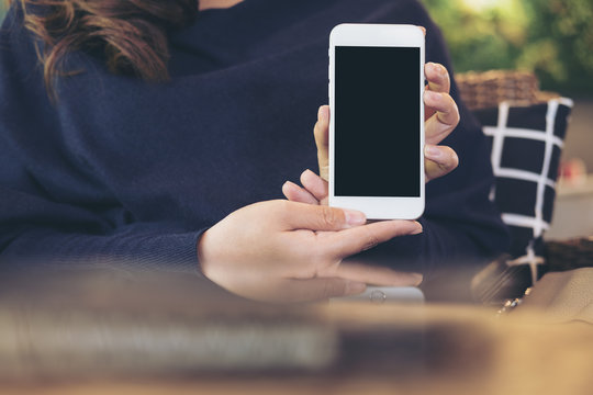 Mockup image of a beautiful woman holding and showing white mobile phone with blank black screen in restaurant