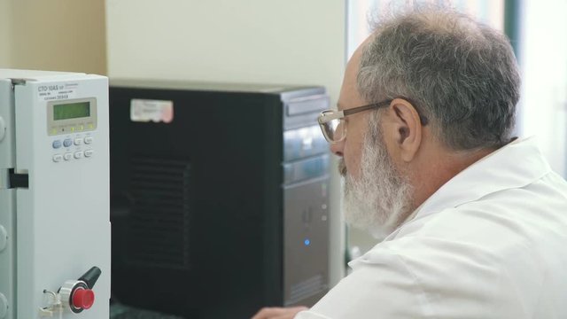 A researcher - a man with glasses and a beard - sits at the computer and watches diagrams on the monitor. Work in the scientific laboratory: the engineer is working on the results of the experiment.