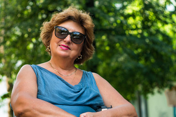 A mature lady in sun glasses. A happy middle-aged woman is smiling while sitting in the park on a sunny day. An outdoor horizontal shot.
