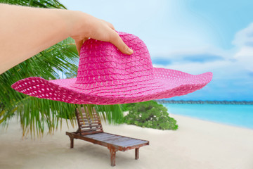 Female hand holding hat and tropical beach with lounger on background. Summer vacation concept