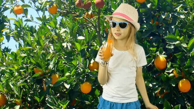 Young caucasian girl drink orange juice in orange garden with orange trees. Green garden summer sunny day and young girl walking in front of trees. Girl dressed in hat t-shirt skirt and sunglasses.