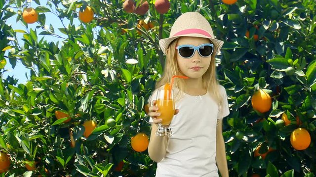 Young caucasian girl turn on and drink orange juice in garden with orange trees. Green garden summer sunny day and young girl standing in front of trees. Girl in hat t-shirt skirt and sunglasses.