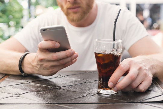 At the cafe. Ginger bearded man is reading smartphone and drinking soda