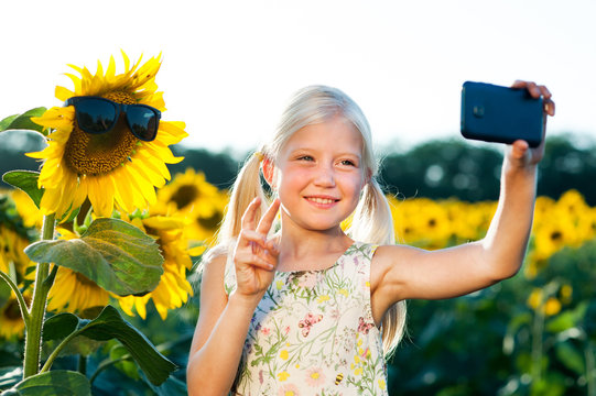 A little beautiful girl is taking pictures of herself on a smart-phone in sunflowers