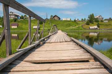 north Russian village Isady. Summer day, Emca river, old cottages on the shore, old wooden bridge and clouds reflections.