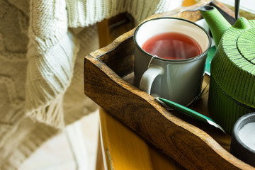 Cup of hot red fruit tea, green pot in tray, candle, knitted sweater hanging on wooden chair, cozy autumn, fall atmosphere, calmness