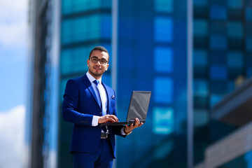 Low angle view of young confident businessman holding laptop and expressing positivity while standing outdoors with office building in the background