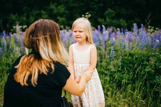 Young mother calming down little crying princess angel daughter. Family at nature. Sad offended child. Young baby blonde girl with long hair standing at ground in wild terrain. Flowers on background.