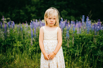 Fototapeta na wymiar Lost, lonely, beautiful child kid little blonde girl with offended, sad, tragic, poor tear-stained expressive emotion face standing in countryside with lupin field on background in wild terrain.