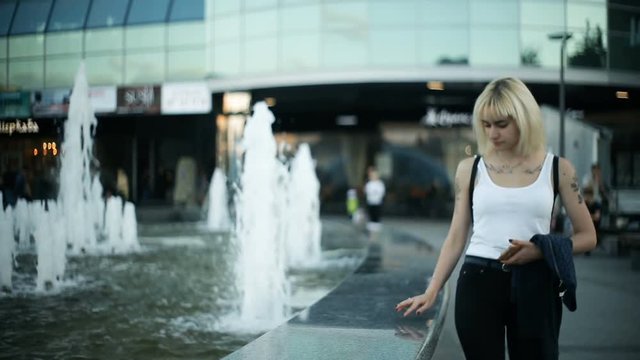A young blonde woman walks in the city, moves towards the camera, passes near the fountain, wets her hands, touches the water.