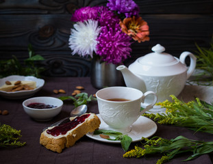 tea in a white cup with slice of bread with butter and jam on it, with white pot, jam, nuts and assorted flowers on a brown wooden background