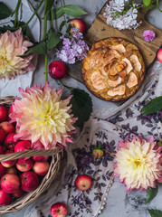 apple pie on wooden desk with a basket of fresh apples,assorted flowers on a grey background, top view