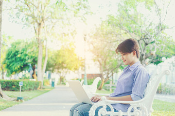 Beautiful short hair woman sitting on white chair under the tree and using laptop for typing outdoor working her business in the park.