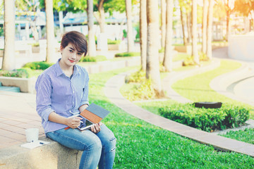 Obraz premium Beautiful short hair woman sitting on the floor and using a tablet for note something and have a favorite beverage in white cup beside between her outdoor working business in the park.