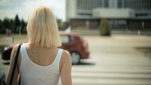 A young blond woman waits for cars to pass, going to cross the road, a red light, a crossing. Cars are passing by. Back view close-up
