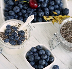 A tasty and healthy breakfast of fresh blueberries, granules, chia seeds, yogurt with blueberries in a glass small jar on a white wooden background.