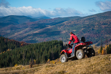 Man riding down on an ATV the hilly road on a background of mountains, forest and blue sky. The concept of an active holiday in the mountains - obrazy, fototapety, plakaty