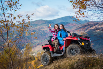 Fototapeta na wymiar Smiling women in jackets on red ATV at the hill makes selfie on the phone with mountains in blurred background