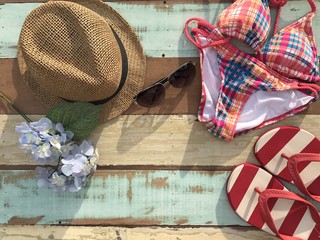 The concept of accessories for summer.Closeup A brown woven hat ,sunglasses,colorful swimsuit and red strip sandals set on pastel wooden board in the sunlight.Copy space with selective focus.