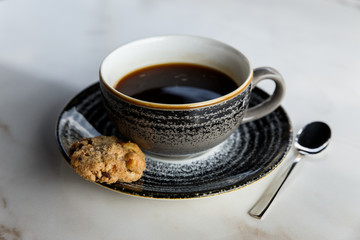 A grey cup of black coffee and a cookie on a saucer, a spoon, on a marble table. Selective focus.