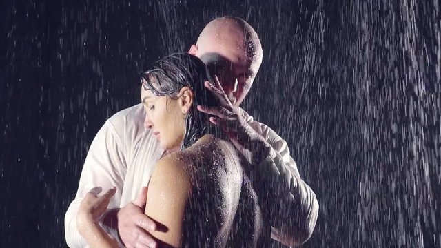 bald man with piercing eyes caresses his woman. they stand in the shower under the streams of water. she's bare back to the camera. he's in a wet white shirt. emotional loving people