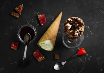 Ice cream in waffle cone with chocolate syrup toppingn in glass on dark black background. Ice cream wafer horns and chocolate syrup on the table, spoon.